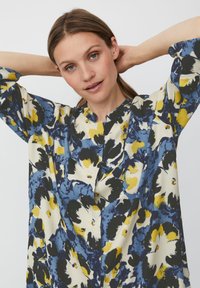 Bloes met bloemenpatroon in blauw, geel en zwarte accenten. Het ontwerp heeft een Mandarin-kraag en driekwartmouwen.