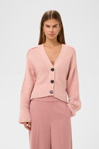 Růžový pletený cardigan s vertikálními žebry, hlubokým výstřihem do V a pěti černými knoflíky. Spárován s odpovídajícími světle růžovými kalhotami.