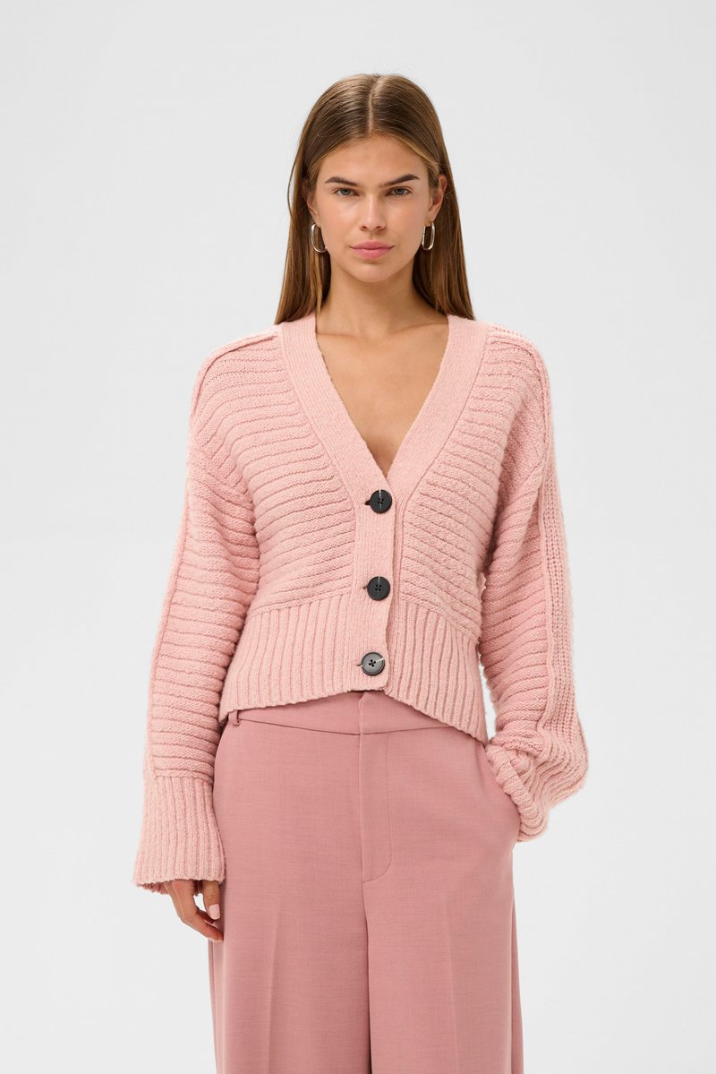 Růžový pletený cardigan s vertikálními žebry, hlubokým výstřihem do V a pěti černými knoflíky. Spárován s odpovídajícími světle růžovými kalhotami.