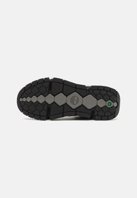 Semelle extérieure en caoutchouc noir avec motif de crampons hexagonaux, comportant un emblème de logo et un accent vert. Texture pour adhérence et durabilité.