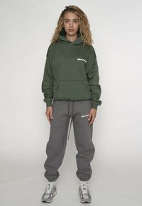 Groene hoodie met een kangoeroezak, spandex manchetten en een wit logo. Gecombineerd met grijze joggingbroek met elastische tailleband en wit logo.