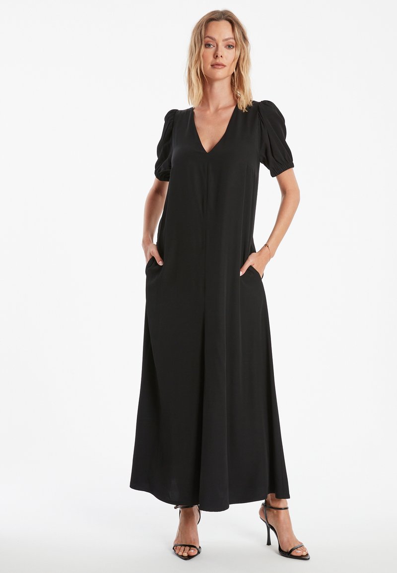 Zwarte maxijurk met een V-hals en puffmouwen. Bevat zijzakken en een soepele, vloeiende stof. Draagt bij strappy zwarte hakken.