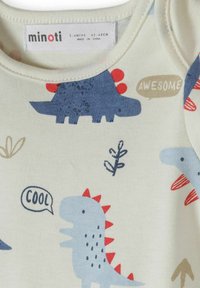 Vêtements pour bébé avec des dinosaures dessin animé bleu et bleu foncé, des pointes rouges et des bulles de dialogue disant "COOL" et "GÉNIAL". Étiquette de taille visible.