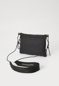 Sac en nylon noir, de forme rectangulaire, doté d'une poche zippée, d'une sangle réglable et d'un branding discret sur la sangle.