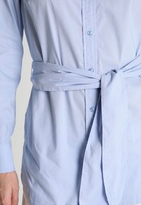 Camisa de manga larga de color azul claro, hecha de algodón, con un cierre de botones en la parte delantera y un cinturón atado en la cintura para un ajuste estilizado.