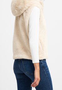Gilet in pelliccia sintetica beige con cappuccio, caratterizzato da una texture morbida. Indossato sopra una maglia bianca a maniche lunghe, abbinato a jeans blu scuro.