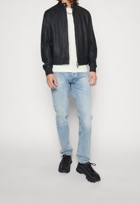 Czarna skórzana kurtka bomberka z prążkowanymi mankietami i kołnierzem, zapinana na zamek, zestawiona z jasnoniebieskimi dżinsami i czarnymi trampkami. Prosty, minimalistyczny krój.