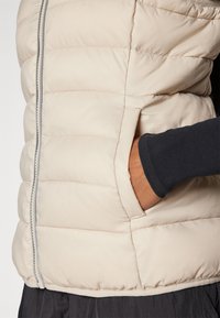 Wattierte Weste in hellem Beige mit gesteppter Textur, Reißverschluss vorne, seitlichen Taschen und taillierter Passform. Lange schwarze Ärmel sichtbar darunter.