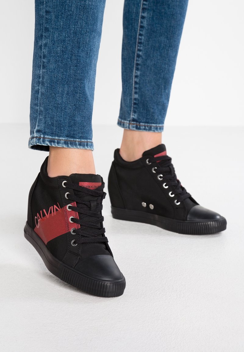 Personne portant des baskets compensées noires avec une bande rouge et le texte « Calvin Klein », associées à un jean denim bleu sur un sol blanc.