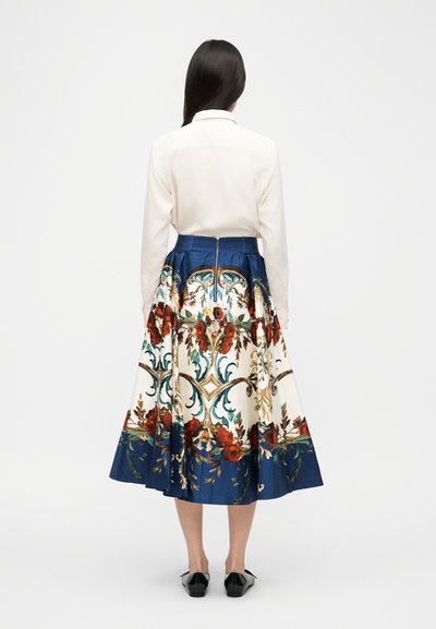 Femme vue de dos portant un chemisier blanc et une jupe midi avec des motifs floraux bleus, rouges et dorés ornés, assortis à des chaussures plates noires.