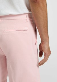 Light pink shorts made of a cotton blend, featuring a rear pocket with a stitched label. Smooth texture with a casual design.