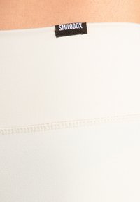 Lichtbeige stof met een gladde textuur, voorzien van een zwart label met de tekst "SMILODOX" en een contrasterende gestikte naaddetail.