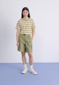 Persona joven con cabello largo y oscuro que lleva una camiseta de rayas verde y beige, pantalones cortos de cargo oliva, calcetines blancos y zapatillas altas color crema.