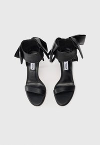 Black leather open-toe heels with ankle straps tied in large bows, viewed from above against a white background.