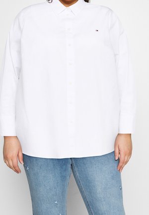 Persona con una camisa blanca abotonada de manga larga y vaqueros rotos de color azul claro, de pie frente a un fondo liso.