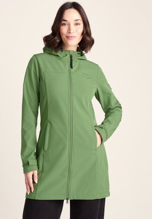 Woman wearing a long green hooded zip-up jacket with front pockets, standing against a plain light background, hands partially in pockets.