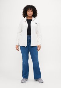 Valge fleece-jakk, millel on lukuga sulgemine, must logo rinnal, kantud musta tipuga, koos siniste laiemate teksade ja hallide tossudega.
