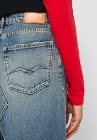 Baklommen på blå Replay-jeans med logo-patch, brukt av en person i en rød langermet skjorte mot en grå bakgrunn.
