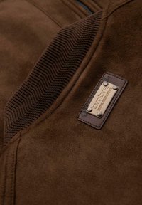 Brown suede jacket with a ribbed collar, featuring a metal brand tag and two studs. Soft texture with a sleek design.