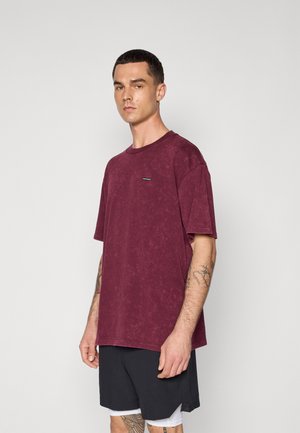 Burgunderfarbens oversized T-Shirt mit rundem Ausschnitt, strukturierter Oberfläche und einem kleinen Logo-Patch auf der Brust. Kombiniert mit schwarzen Sportshorts.
