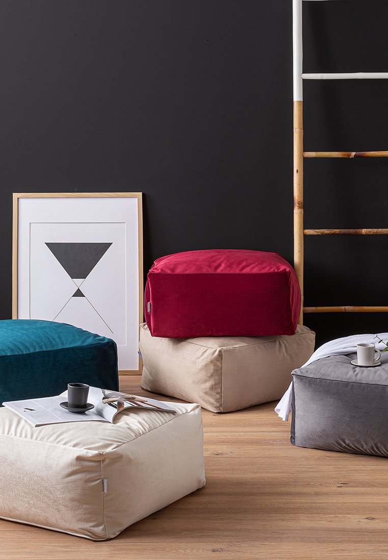 Tre sacchi per i fagioli di diversi colori: turchese, rosso e beige, realizzati in tessuto morbido con texture lisce, posizionati su un pavimento in legno vicino a opere d'arte.