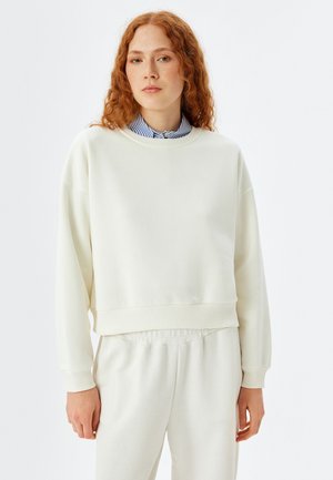 Cremfarbener Pullover aus weichem Material, mit rundem Halsausschnitt, überschnittenen Schultern und ribbelten Bündchen. Kombiniert mit passenden hellen Hosen.