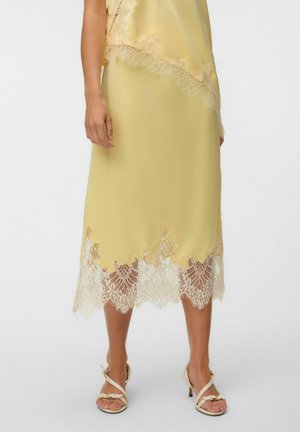 Jupe midi en satin jaune avec bordure en dentelle blanche à l'ourlet, portée avec des sandales à talons beiges à brides sur fond clair.
