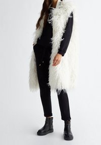 Weißer, flauschiger ärmelloser Mantel über einem schwarzen Pullover und einer schwarzen Hose, ergänzt durch schwarze Stiefeletten. Der Mantel hat ein strukturiertes, fransiges Aussehen.