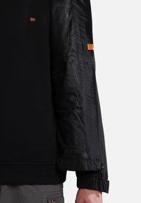 Sort jakke med strikket overdelen, ærmer i mesh og en orange detalje. Inkluderer lynlåse ved ærmekanterne og et lille rødt og hvidt emblem.