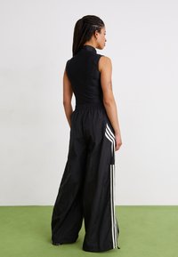 Kvinne med flettet hår står vendt mot høyre iført ermeløs svart topp og vide sorte bukser med hvite striper på et grønt gulv.