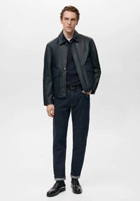 Schwarze Lederjacke mit Kragen, Druckknopfverschluss und zwei vorderen Taschen, kombiniert mit dunklen Jeans und schwarzen Schuhen.