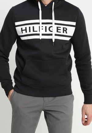 Czarna bluza z kapturem z białymi sznurkami i białym prostokątnym paskiem na piersiach z napisem „HILFIGER”, noszona z szarymi spodniami.
