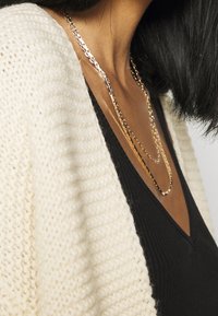 Layered gold chains rest over a black v-neck top, complemented by a textured cream cardigan with a wide collar and knitted pattern.