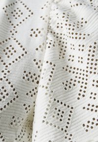 Weißer Stoff mit einer strukturierten Oberfläche, die bestickte Diamantformen und kleine Aussparungen aufweist. Das Muster ist repetitiv und geometrisch.