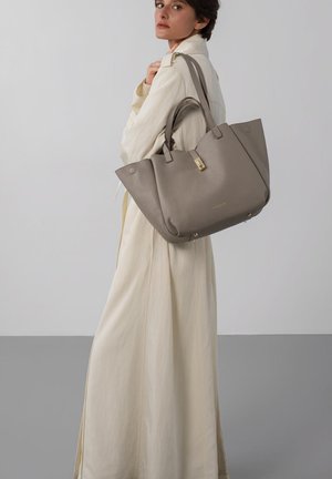 Femme portant un long trench-coat beige et un grand sac fourre-tout en cuir taupe avec une fermeture dorée sur l'épaule, sur fond uni.