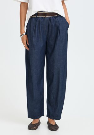 Personne portant un pantalon large taille haute bleu marine avec des plis, une ceinture marron, un haut blanc et des chaussures tissées sombres sans lacets, debout les mains dans les poches.