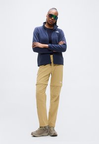 Ζακέτα φλις με φερμουάρ σε ναυτικό μπλε, χακί παντελόνι cargo και γκρι μποτάκια πεζοπορίας. Γυαλιά ηλίου με πολύχρωμους φακούς. Λειτουργική εξωτερική ένδυση.