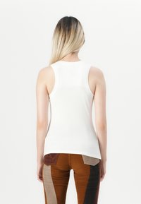 Hvit ribbestrikket tanktopp med rund hals og ermeløs design, kombinert med brune kordfløyelsbukser med paneler i forskjellige farger.