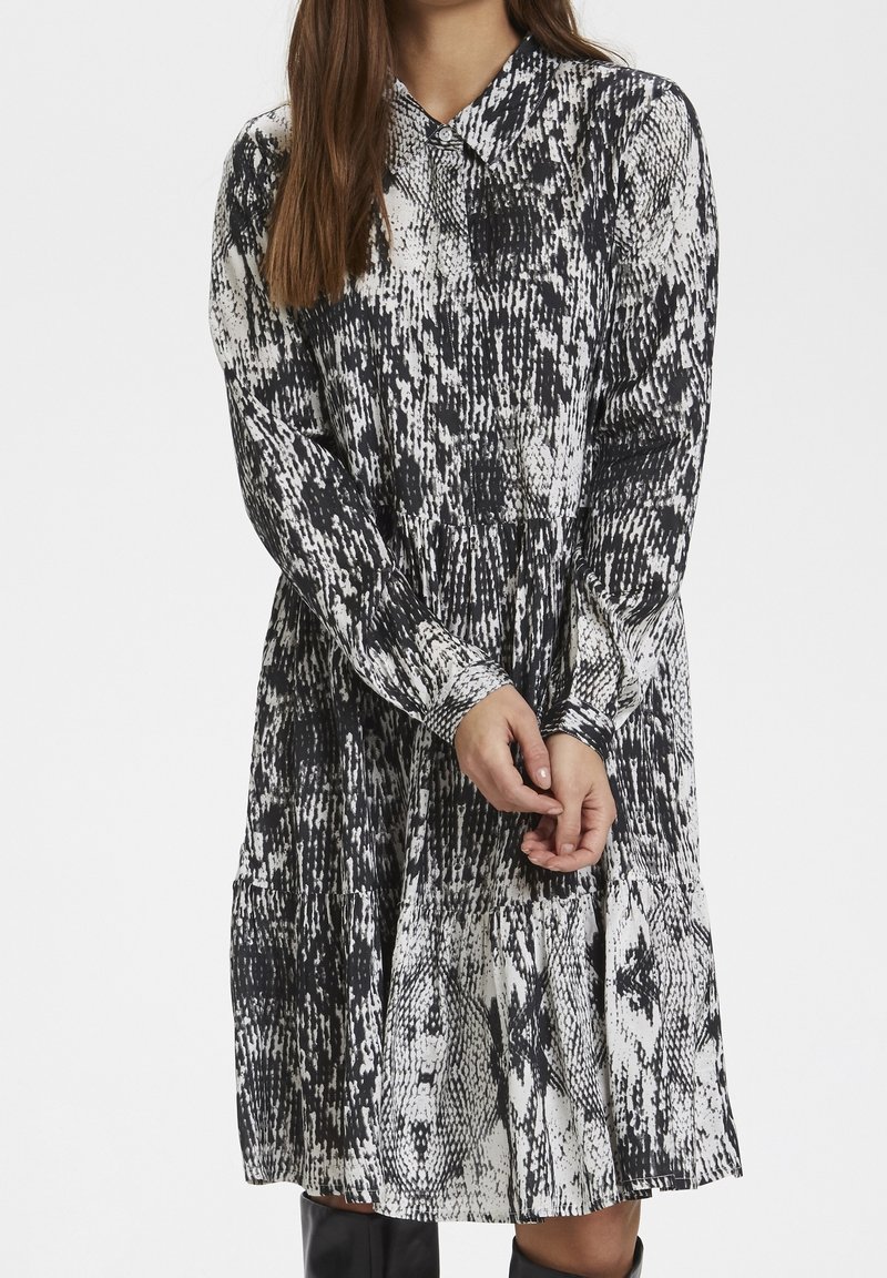 Robe à motifs noir et blanc avec col à revers, manches longues et partie jupe plissée, en tissu léger.