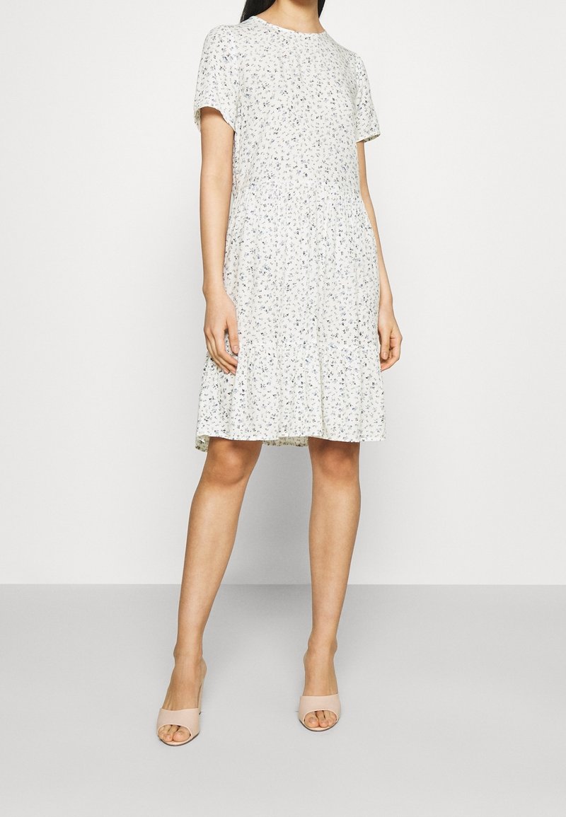 Femme portant une robe blanche mi-longue à manches courtes avec un petit motif floral, portée avec des talons ouverts beiges sur un fond uni.