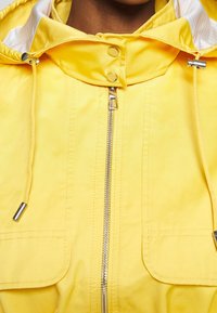 Veste jaune avec fermeture éclair argentée, boutons-pression, cordons avec embouts métalliques et capuche doublée de tissu blanc.