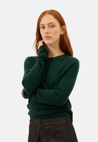 Dunkelgrüner Strickpullover mit langen Ärmeln und rundem Ausschnitt, mit lockerer Passform und gerippten Bündchen, kombiniert mit dunklen Jeanshosen.