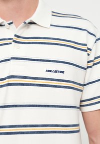 Πόλο μπλούζα σε λευκό με οριζόντιες ρίγες σε ναυτικό μπλε, ανοιχτό μπλε και κίτρινο. Διαθέτει γιακά και κεντημένο λογότυπο «Hollister» στο στήθος.