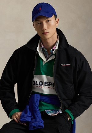 Black jacket with a collar, layered over a white and green Polo Sport shirt. Blue cap and knitted blue sweater tied around the waist, plaid shirt underneath.
