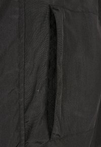 Chaqueta de tela negra con un bolsillo lateral con cremallera. El material tiene una textura suave y parece ligero, con mínimas arrugas.