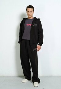 Junger Mann in einem schwarzen ZIP-Hoodie, schwarzen Jogginghosen mit dem Text "Local Heroes", einem dunkelgrauen "777"-T-Shirt und weißen Turnschuhen, der drinnen steht.