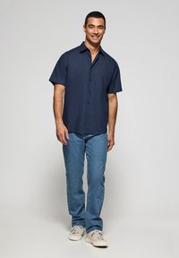 Camisa de botones de manga corta en color azul marino con un bolsillo en el pecho, combinada con jeans de tonos azules claros y zapatillas crema, presentada sobre un fondo neutro.