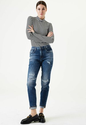 Haut à col roulé rayé, jeans en denim avec des patches déchirés, et mocassins noirs avec des détails en or. Denim à lavage clair, poignets retroussés.