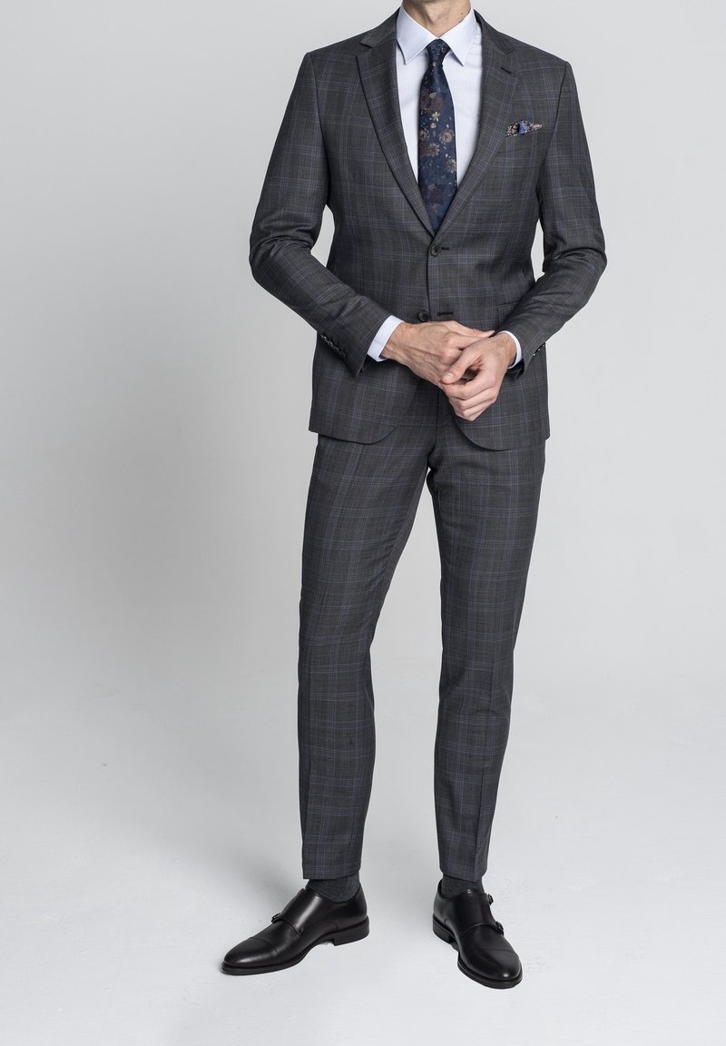 Homme portant un costume gris foncé à carreaux avec une chemise blanche, une cravate à motifs floraux, une pochette et des chaussures habillées noires sur un fond uni.