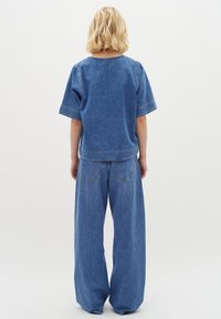 Denim ruházat, amely egy laza felsőt és bő szárú nadrágot tartalmaz. Kék anyag, látható varrási részletekkel, struktúrált varratokkal és két hátsó zsebbels.